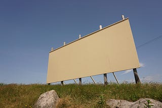 A blank, white billboard on the side of a deserted highway with some dry rocks and sun damaged grass.