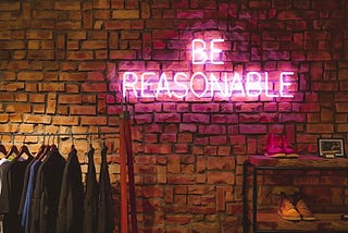 A picture of the words “Be Reasonable in neon lights against a red brick wall, a row of jackets on a rack and a table with a pair of trainers feature below