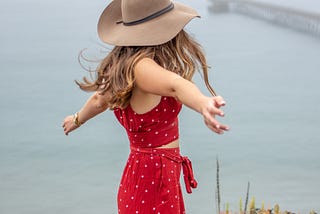 A woman in a red dress stands on a cliff edge with her arms outstretched