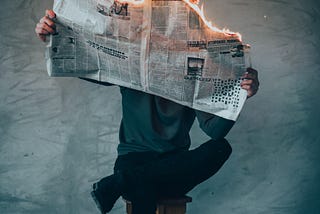 A person leaning against a wall reading a newspaper. The paper is on fire.