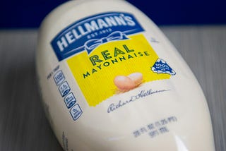 A bottle of Hellmann’s Mayonaise lays on a gray, wooden table. The label reads: “Hellmann’s Est. 1913, Real Mayonnaise.”