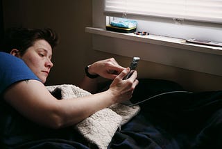 A woman is lying in bed, scrolling through her phone. The surroundings are barren, with just a few items on a windowsill.
