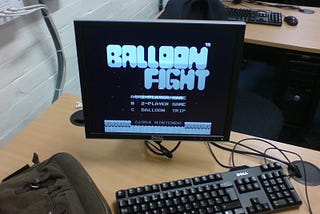 A computer screen on a desk with keyboard and mouse showing the title screen of the game “Balloon Fight” in black and white.