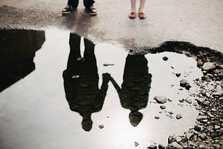 a couple holding hands looking at their reflection in a puddle.