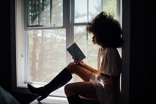 A young black women sitting by the window reading a book.