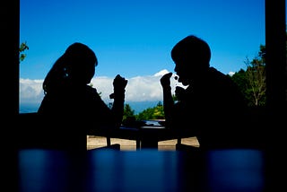 A silhouette of two people sitting at a table against a bright blue sky with clouds.