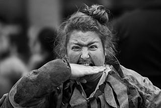 person with a mustache and long hair in bun appears to bite their own hand performatively