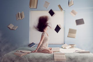 Woman sitting on bed covered with books whips her hair back. Open books float in the air around her.