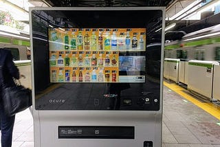 Vending machine at train station, with trains zooming by in both directions. A busnessman walks by the side of the vending machine.