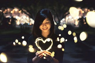 A woman smiles as she looks at lights shaped as a heart.
