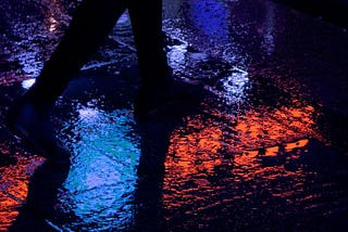 A man solemnly walks in the rain by a tea bar.