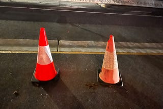 two traffic cones, one damaged, below words “leave everything unfinished so that humanity might creep in”
