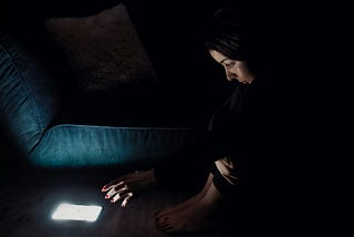 Woman sitting on the floor in the dark reaching for her glowing phone.