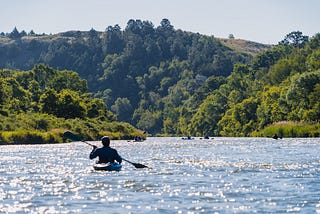 Photo of a man in a kayak paddling in water. Green hills are in the distance.