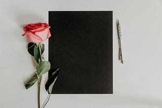 A pink rose, blank black page, and pen representing the creative process of crafting personalized poetry with a Poetry Generator.