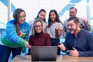 Men and women gathered at a table staring at a laptop while laughing and cheering. The corporate joy is palpable. One woman is wearing a Microsoft Vista themed sweater.