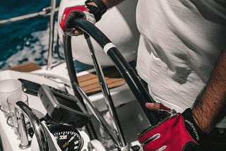 Red gloved hands of a hairy-armed man griping the helm on a boat with his course holding steady.