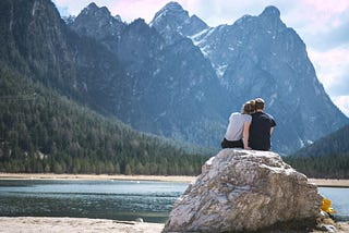 A couple sitting on a rock facing a lake with trees and mountains beyond the lake in the background.