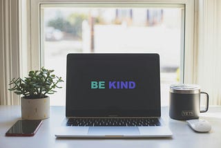 Laptop next to a window, plant, and mug. The computer screen says “be kind.”