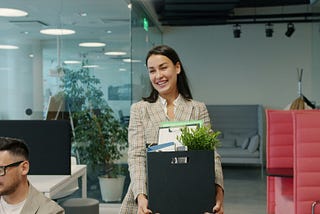 Happy young lady former employee is quitting job leaving office room with box of things walking and talking to ex-colleagues. People and employment concept.