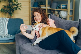 Image of woman sitting with her dog on a sofa taking a selfie.