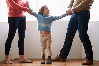 Mother holding one arm of a little girl, and father holding one arm, both appearing to tug slightly.