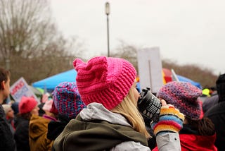 A blonde woman wearing a pink hat drinking a coffee, at the women’s march.
