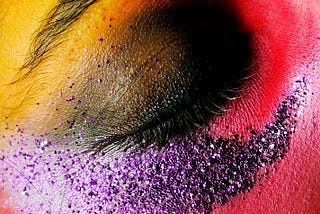 A picture of a closed eye with a painted black eyelid surrounded by dark pink, purple glitter, and yellow around the eyebrow.