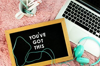 Aerial view of pink plushy carpet. Coffee mug with wooden pencils, an open Macbook, teal over-ear headphones, and a letter board with the phrase, “You’ve got this.”