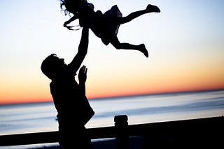 Along a wooden dock fence in the forefront of a distant orange sunset mirroring the dark, thin horizon on the water’s edge are the silhouettes of a Father holding his daughter high in the air with one hand. The daughter’s long hair is scattered around in a web of delight, and her trusting posture reveals how often her Father has lifted her up in the air like this. The joy and connection they share are evident by the level of trust with such a maneuver of grace.