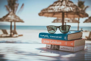 Sunglasses sitting on a stack of three books on a table with a beach in the background.