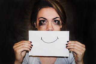 Woman with smudged makeup holding a piece of paper with a fake smile in front of her face.