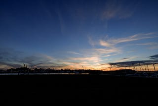 Large industrial cranes on the former Mare Island Naval Base in Vallejo, Calif. are silhouetted against the sky at sunset. The Napa river is visible in the foreground. On the right of the image are the masts of several docked sailboats.