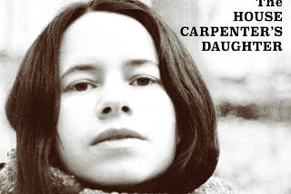 The cover art of Natalie Merchant’s ‘The House Carpenter’s Daughter’ album shows her looking at the camera. The photo is in black and white. The title is in the upper right-hand corner in black type and block lettering. Merchant’s name appears in a banner across the top with a light blue background. The words ‘a collection of tradional & contemporary folk music’ run across the bootom in white type against a black background.