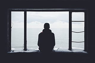 a lone man, sitting on a windowsill, facing the ocean, seen from behind.