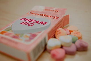 Box of Sweethearts candies with a heart-shaped candy reading ‘DREAM BIG’ in bold red text, surrounded by colorful pastel heart-shaped candies spilling out by Kenny Eliason