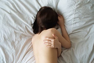 A brunette in a rumpled bed is hiding her face and has one arm wrapped around her body.