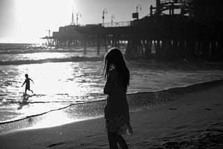 A grayscale image of a woman standing on the shore at the beach. There is also a  silhouette of a child playing in the water.