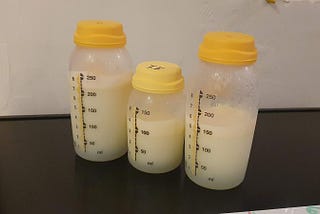 A photo of three milk bottles with varying amounts of milk in them, the size of two of the bottles is 250ml and the other bottle is 150ml, all the bottles have yellow lids and they have measurement markings up their side, the milk inside them has separated slightly with thicker cream on the top and greener/yellowy water near the bottom