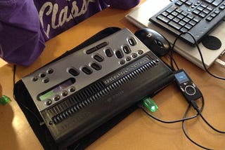 A blind teenager sits at a desk in front of a braille notetaker, a desktop computer and a MP3 player.