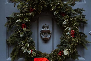 A Christmas garland hangs on a grey front door, circling a metal door knocker
