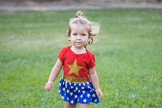 A cute little girl dressed as a superhero walking across green grass towards you, the viewer.