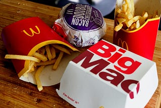 A packaged fast-food meal on a wooden table, with fries, a burger, and a Big Mac box. A familiar, ready-made combination designed for speed, convenience, and immediate satisfaction rather than attention or presence.