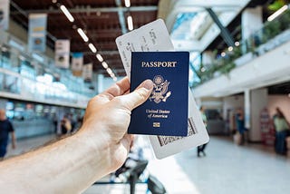 person in an airport holds up a blue US passport and a plane ticket