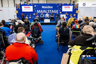A photograph of the audience and a panel of speakers on the main stage at Naidex. Many of the audience and speakers are wheelchair users.