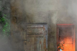 The front of a small old house shrouded in smoke, fire light seen through the window.