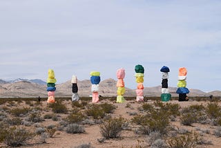 Colorful boulders stacked in seven towers are set against a desert landscape with sparse vegetation and distant mountains under a cloudy sky. These seven towers depict The Seven Components of Authentic A.S.S.™ Infrastructure for coaches, freelancers, and solopreneurs who want to fix their failing systems and achieve business success. Image credit: Lyle Hastie (Unsplash) — “7 rock-based structures in desert field” under the following license: Unsplash Free to Use.