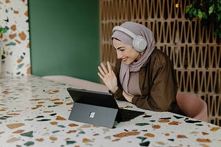 A woman wearing headphones and a headscarf is sitting at a bench and waving at someone having a video chat with her, via her tablet computer.