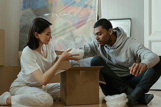 A man and a woman sitting down on the floor while getting things out of a box.