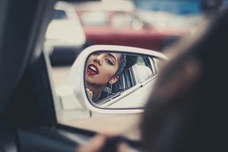Woman putting on red lipstick in the side mirror of a car